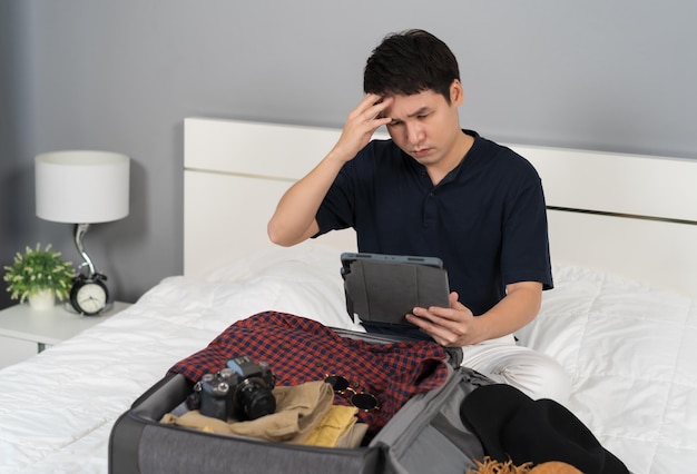 Photo stressed man using tablet and packing clothes into suitcase on a bed holiday travel concept