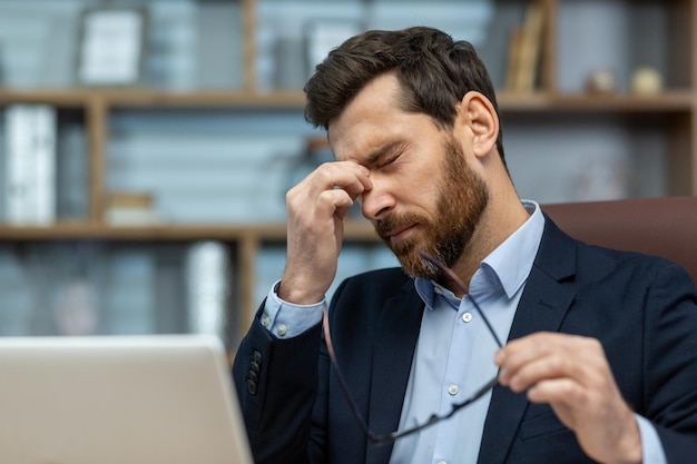 stressed mature businessman rubbing eyes while working on laptop in a classic office