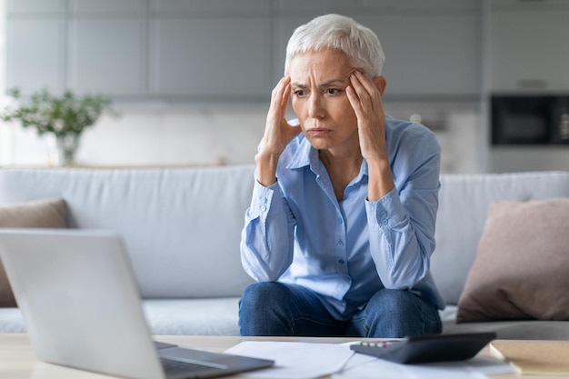 Photo stressed mature woman touching head in despair calculating budget indoors