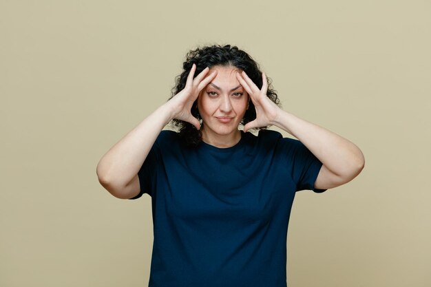 Stressed middleaged woman wearing tshirt keeping hands on head looking at camera isolated on olive green background