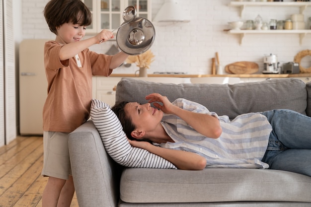 Stressed mother try to sleep on sofa suffer from headache from naughty son bang metal kitchenware