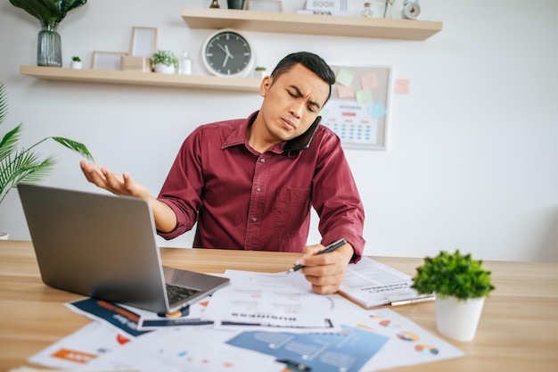 Stressed out, a man works in the office with his laptop, papers on the desk and a phone.