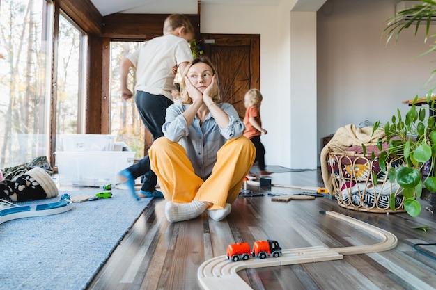 Stressed out mother sitting in middle of toys while children naughty running around her at room