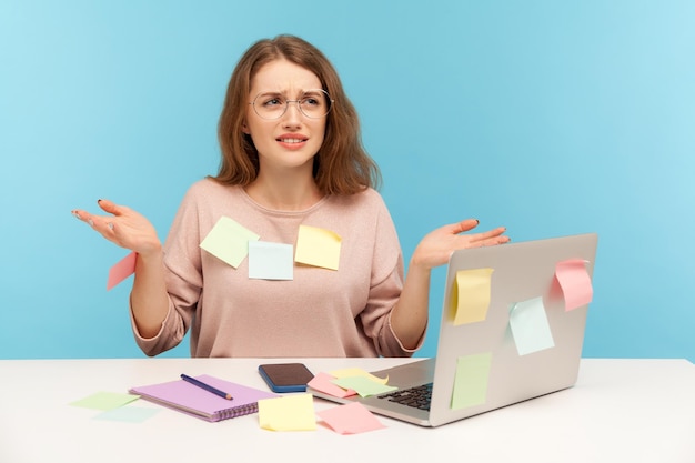 Stressed overworked woman in nerd glasses sitting covered with sticky notes, looking outraged by workload at home, raising hands in indignant gesture. indoor studio shot isolated on blue background