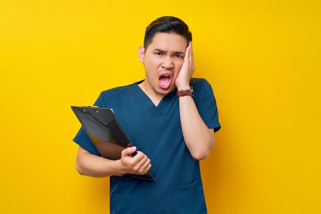 Stressed professional young asian male doctor or nurse wearing a blue uniform holding clipboard and touching his head with hand isolated on yellow background healthcare medicine concept