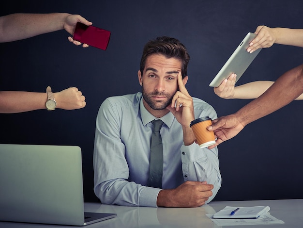 Stressed and strained from work Portrait of a young corporate businessman sitting at a desk surrounded by hands reaching in with office items