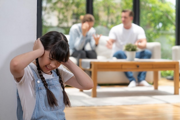 Stressed and unhappy girl huddle in corner cover her ears with painful expression while her parent arguing in background Domestic violence and traumatic childhood develop to depression Synchronos