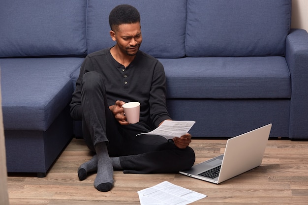 Stressed upset man sitting on floor holding rose cup with drink and document in both hands, reading attentively, using his laptop