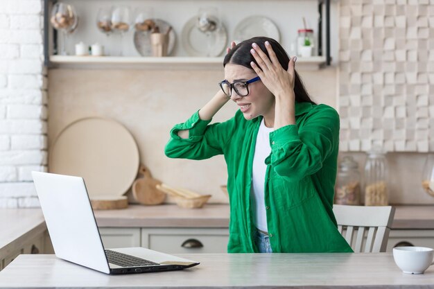 Stressed woman at home working in kitchen using laptop got bad news online brunette holding her head