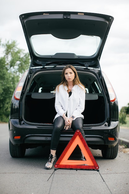 壊れた車を見て、側面図で田舎道でエンジンをチェックしているストレスの多い女性