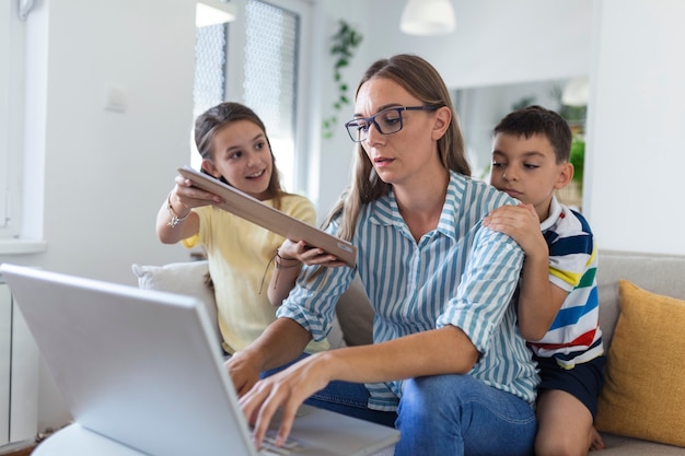 집에서 일하는 아이들과 함께 스트레스를 받는 여성.피곤한 젊은 어머니는 소파에 앉아 노트북과 문서 작업을 하는 반면 어린 아이들은 즐겁게 놀고 소음을 냅니다.