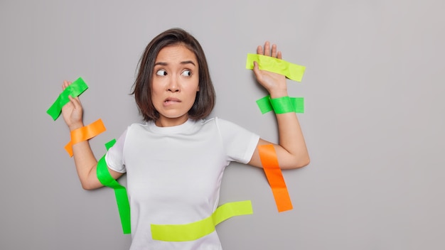 Stressed worried Asian woman bites lips dressed in casual white t shirt stuck to grey wall with colorful adhesive tapes looks away poses against grey wall