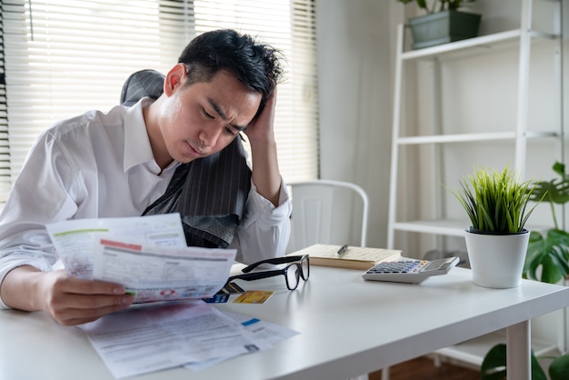 Stressed young asian businessman holding so many expenses bills such as electricity bill,water bill,internet bill,cell phone bill and credit card bill in his hand no money to pay debt