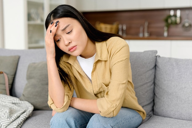 stressed young asian woman touching head and feels dizzy suffering from headache and migraine pain sits on the sofa at home