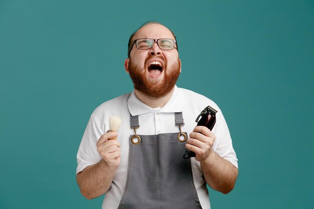 Stressed young barber wearing uniform and glasses holding shaving brush and hair trimmer screaming with eyes closed isolated on blue background