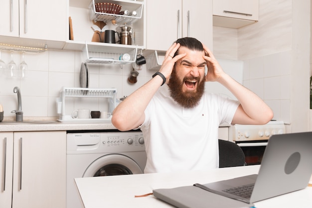 Stressed young bearded man is having a headache while working from home at the laptop.