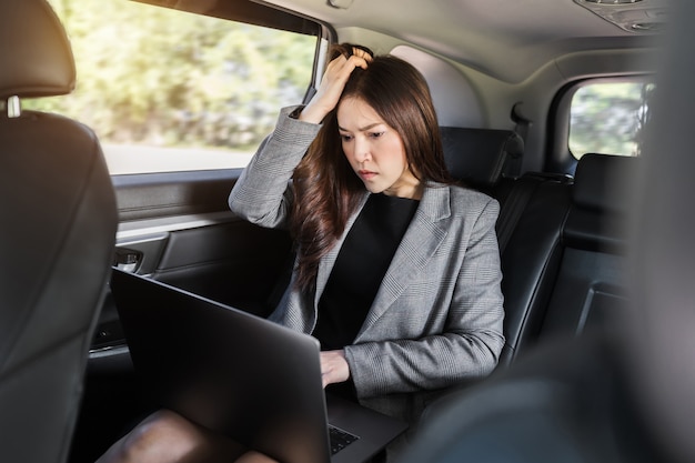 Stressed young business woman using laptop computer while sitting in the back seat of car