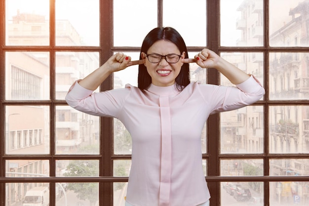 Stressed young businesswoman covering ears with fingers trying not to hear checkered window with str