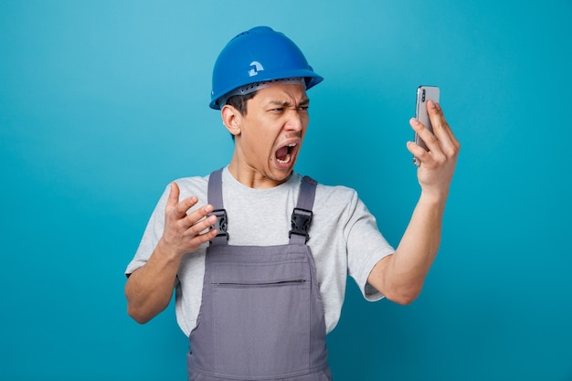 Foto ha sottolineato il giovane operaio edile che indossa il casco di sicurezza e la tenuta uniforme e guardando il telefono cellulare urlando tenendo la mano in aria