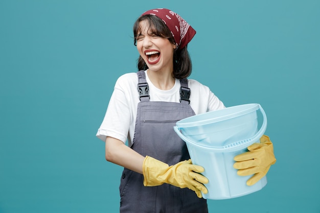 Stressed young female cleaner wearing uniform bandana and rubber gloves holding bucket with both hands screaming with closed eyes isolated on blue background