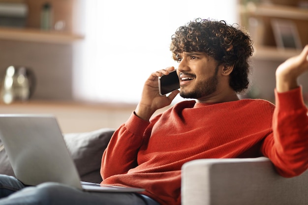 Stressed young indian man talking on cellphone and using laptop at home