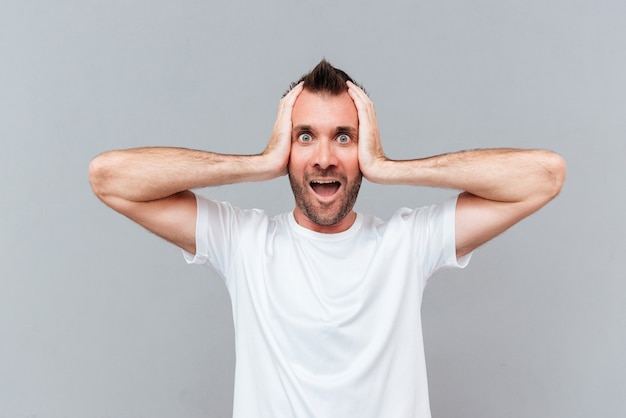 Stressed young man covers ears by hands and shouting over gray wall