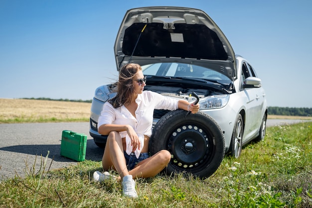 彼女の車のエンジンを見て強調した若い女性。ロードトリップの問題。車は修理が必要です。