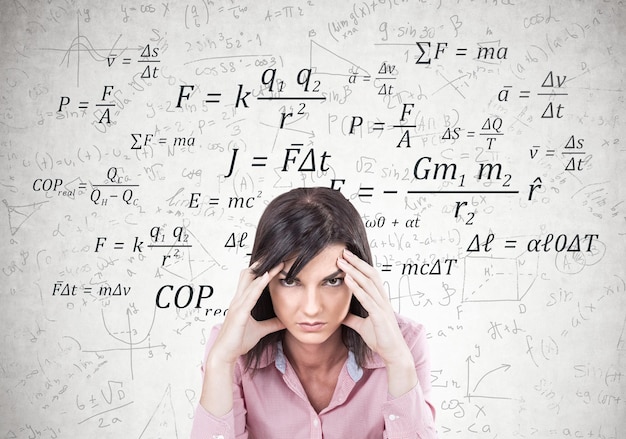 Stressed young woman wearing a pink shirt and sitting with her fingers touching her temples. A headache. A concrete wall with formula on it