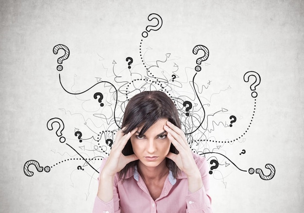 stressed young woman wearing a pink shirt and sitting with her fingers touching her temples. a headache. a concrete wall with question marks
