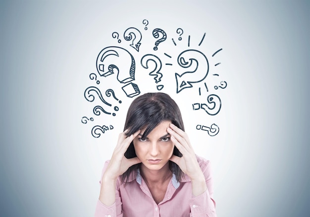 stressed young woman wearing a pink shirt and sitting with her fingers touching her temples. a headache. a gray wall with question marks.
