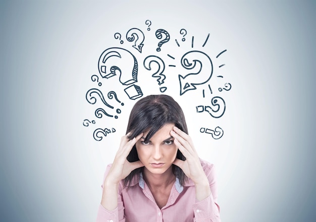 Stressed young woman wearing a pink shirt and sitting with her fingers touching her temples. A headache. A gray wall with question marks.