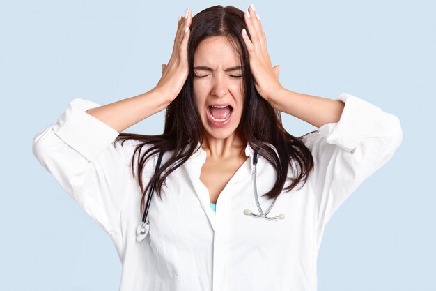 ストレスの多い女性医師または専門家が頭に手をつないで、ひどい頭痛に苦しみ、痛みから叫び、口を広く開いたままにし、頭に傷を付け、青い壁に分離した白いローブを着ています。