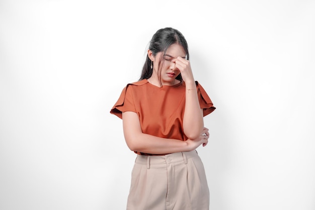 Stressful young Asian woman in a brown shirt and taupe pants is holding her head in her hands She is in pain or discomfort
