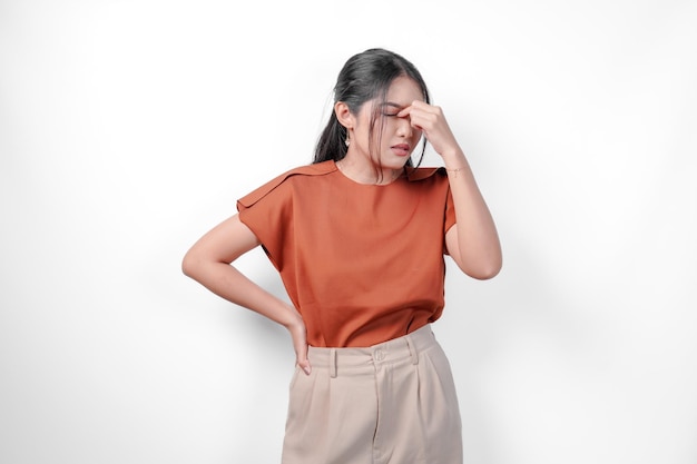 Stressful young Asian woman in a brown shirt and taupe pants is holding her head in her hands She is in pain or discomfort