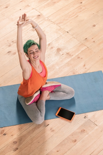 Stretching body. Cheerful green-haired businesswoman stretching her body sitting on sport mat