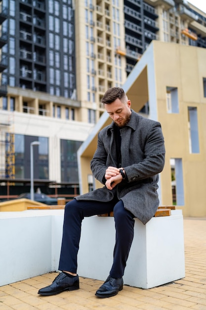 Strict adult man sitting on bench on modern contemporary street and watching on his wirstwatch.