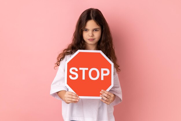 Strict bossy adorable little girl holding red stop sign looking at camera with serious expression