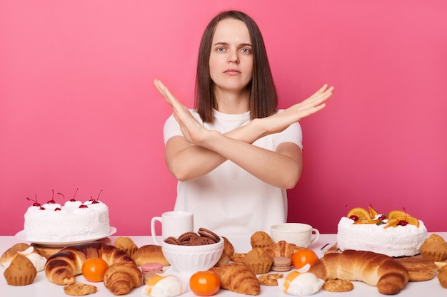 Foto rigorosa donna dai capelli castani in maglietta bianca seduta a tavola con pasticceria che non mostra alcun gesto che incrocia le mani smette di mangiare dolci isolati su sfondo rosa