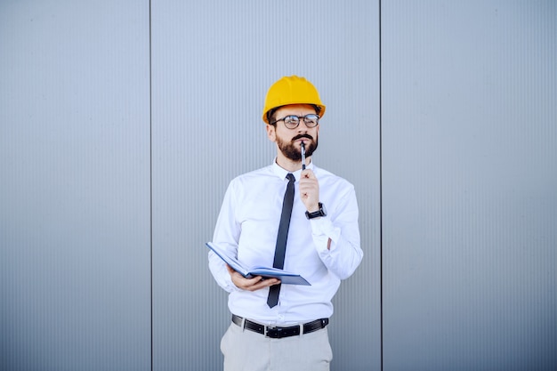 Strict caucasian director in shirt and tie, with eyeglasses and helmet on head standing