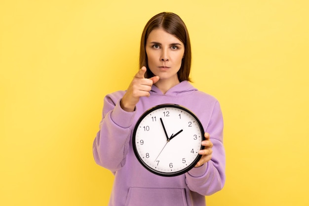 Strict dark haired young adult woman holding wall clock in hands and pointing to camera at you, time management, you are late, wearing purple hoodie. Indoor studio shot isolated on yellow background.