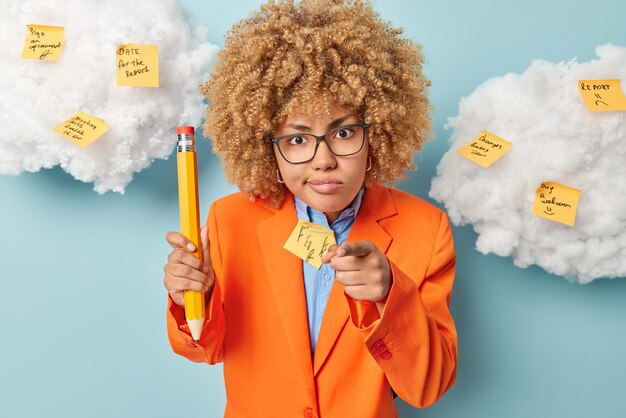Strict female teacher points index finger forward blames you
holds big wooden pencil asks pupils not to make noise dressed in
formal orange jacket isolated over blue background uses
stickers