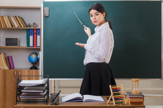 strict looking at camera young female teacher standing in front blackboard and points at blackboard with pointer in classroom