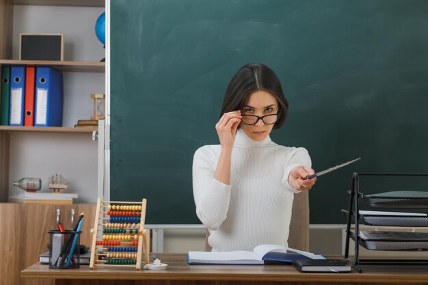 strict looking at camera young female teacher wearing glasses holding pointer sitting at desk with school tools on in classroom