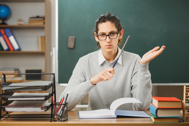 strict looking at camera young male teacher wearing glasses holding pointer sitting at desk with school tools on in classroom