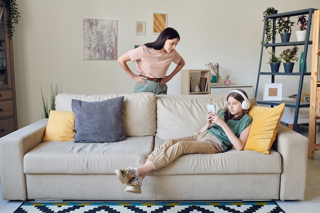 Strict mother keeping her hands on waist while standing by\
couch where her lazy teenage daughter with smartphone listening to\
music