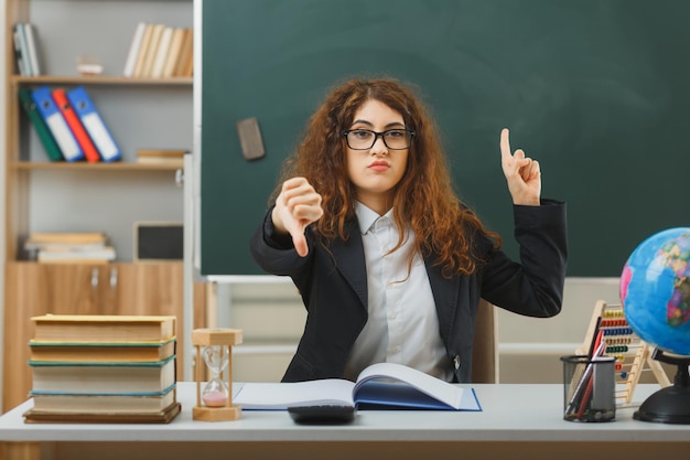 strict points at up showing thumbs down young female teacher wearing glasses sitting at desk with school tools in classroom