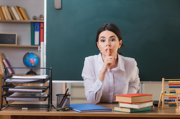 教室で学校の道具を持って机に座っている若い女性教師が沈黙のジェスチャーを厳密に示す