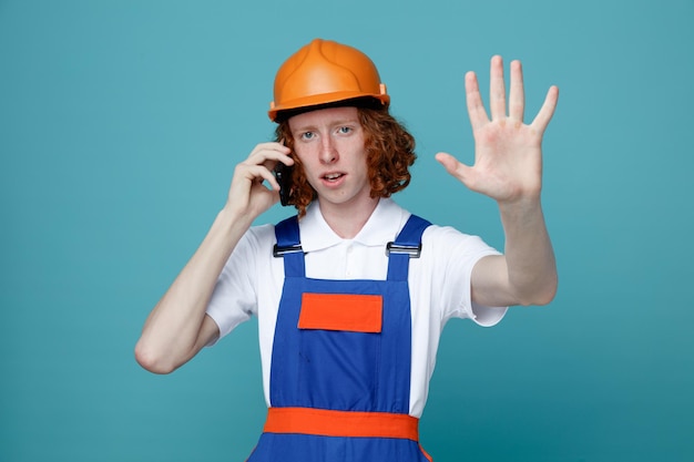 Strict showing stop gesture young builder man in uniform speak on the phone isolated on blue background