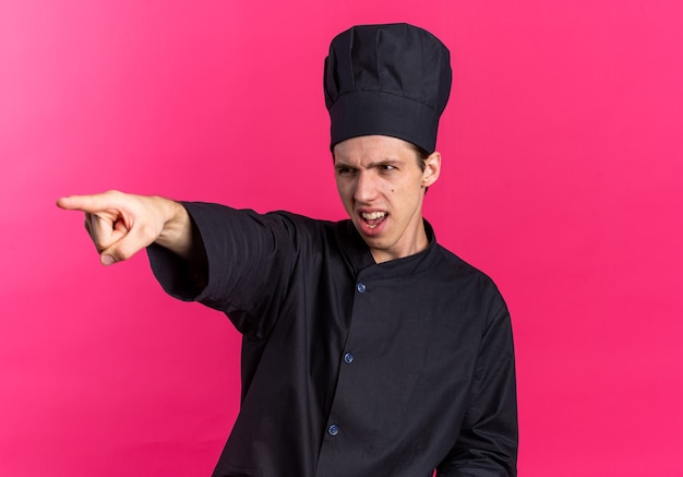 Strict young blonde male cook in chef uniform and cap looking and pointing at side isolated on pink wall