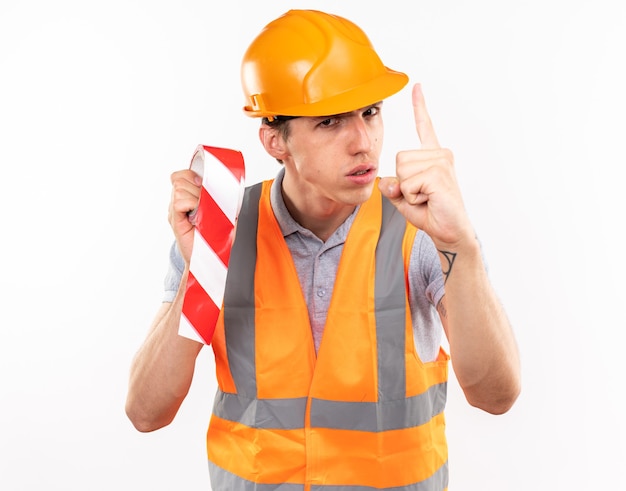 Strict young builder man in uniform holding duct tape showing one 